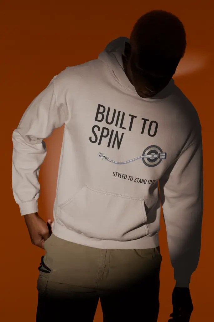 Person wearing a light heather grey Built to Spin unisex hoodie with black text graphic, under dramatic lighting with an orange background.